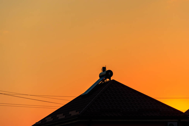 Napenergiás vízmelegítő egy lakóház tetején naplementekor. Megújuló energia háztartás céljára - Fotó, kép