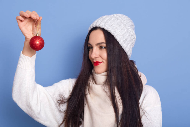 Brunette woman in white cap and sweater, holding red Christmas ball in hands and looking at it, girl with red lips posing isolated over blue background, female being ready to decorate home for holiday - Φωτογραφία, εικόνα