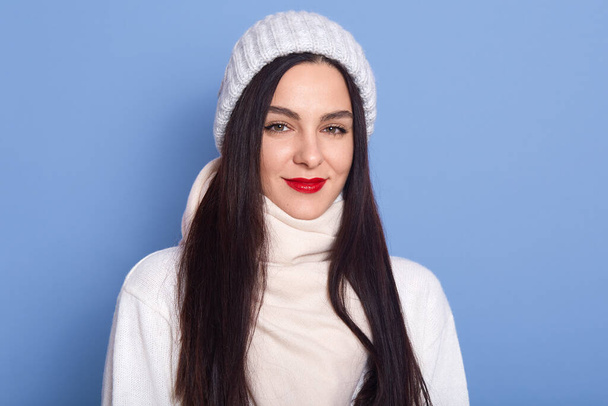 Woman feeling cold, wearing white cap, sweater and scarf, looking directly directly at camera with charming smile and red lips, girl with long dark hair posing isolated over blue background. - Foto, afbeelding
