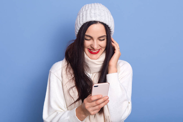 Portrait of nice attractive satisfied cheerful girl holding in hands and using cell phone, reads happy news, lady stands isolated on blue background, female wearing white pullover, scarf and cap. - Foto, afbeelding