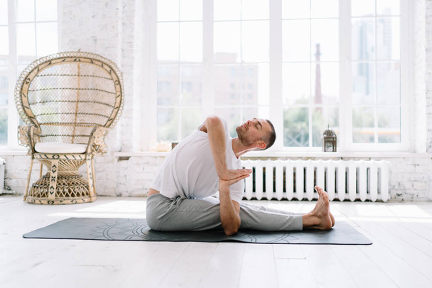 Calm handsome male with closed eyes sitting on carpet in asana recreating and feeling body muscles, pensive hipster guy keeping healthy lifestyle recreating during yoga training at home interior - Фото, изображение