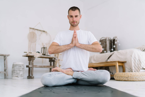 Portrait of handsome male in lotus pose holding hands in namaste enjoying spiritual meditation at flat, hipster guy looking at camera during morning yoga practice in modern interior apartment - Valokuva, kuva