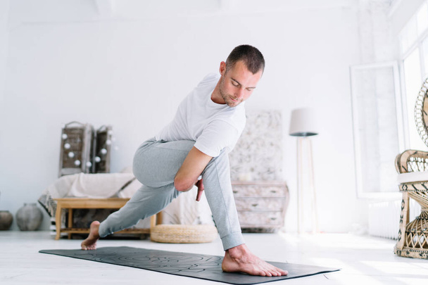 Serious pensive caucasian male in casual wear standing on carpet in asana recreating and breathing, calm hipster guy keeping perfect body shape on morning yoga training at cozy home interior - Zdjęcie, obraz