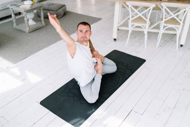 Concentrated calm caucasian man in casual wear sitting in asana enjoying healthy mind and body at home training, positive young male satisfied with yoga and meditation doing exercises on carpet - Zdjęcie, obraz