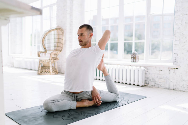 Handsome man in shirt with copy space for brand name or label practicing yoga at home interior, concentrated calm male sitting in asana breathing and stretching body for healthy lifestyle - Photo, image