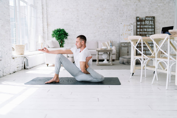 Concentrated caucasian male in casual wear sitting in asna on carpet feeling recreation and relaxing,handsome hipster guy enjoying morning yoga practice at home interior feeling spiritual strength - Foto, afbeelding
