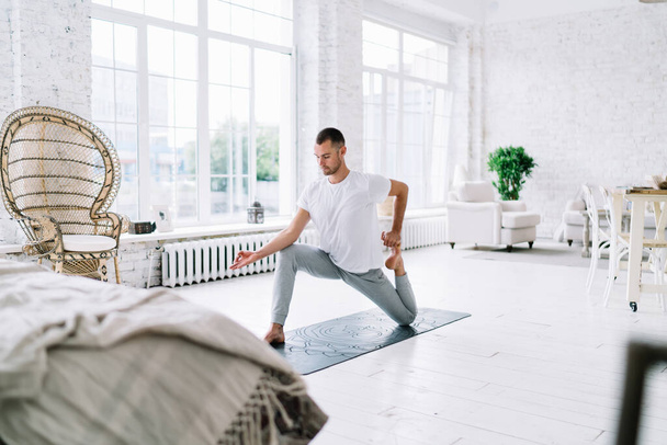Pensive caucasian male in casual wear standing in asana on carpet in apartment feeling balance and spirituality for healthy mind, hipster guy enjoying yoga pose on training at home interior on leisure - Fotografie, Obrázek