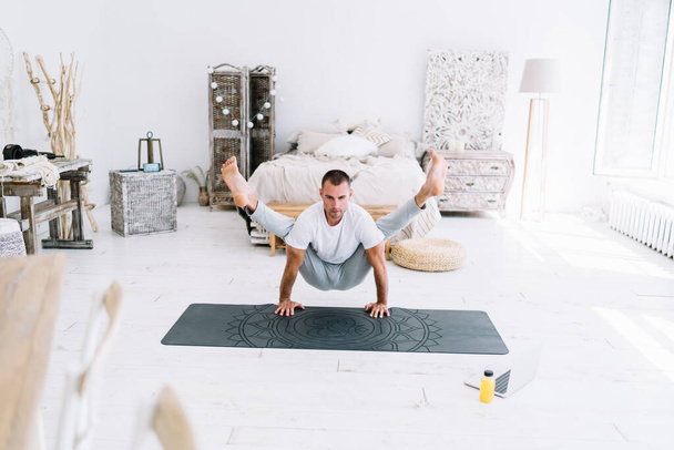 Smiling hipster guy in casual wear having morning yoga training at home interior using laptop computer for online lesson with instructor, confident strong man standing in asana breathing and rest - Photo, Image