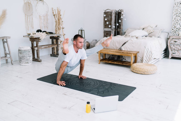 Determined strong hipster guy in casual wear training body and mindfulness at home interior in morning, 20s serious caucasian guy standing in asana keeping perfect body shape and mindfulness - Фото, зображення