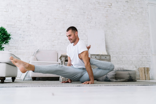 Smiling hipster guy in casual wear sitting in asana stretching muscles on morning training enjoying healthy lifestyle, handsome positive man recreating and training staying at home in apartment - Fotó, kép