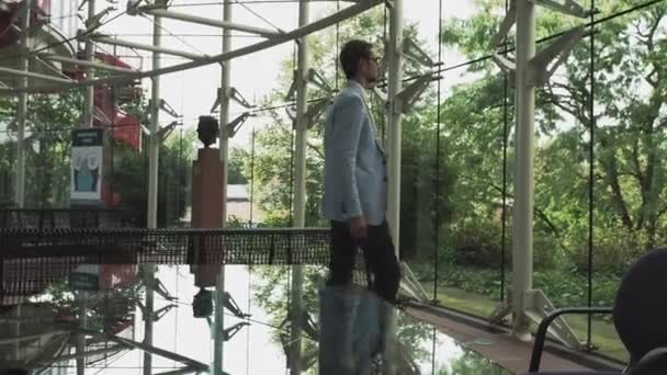 Um jovem bonitão em um terno de arquiteto ou advogado anda em torno de um escritório moderno. Reflexão em uma mesa de vidro. - Filmagem, Vídeo