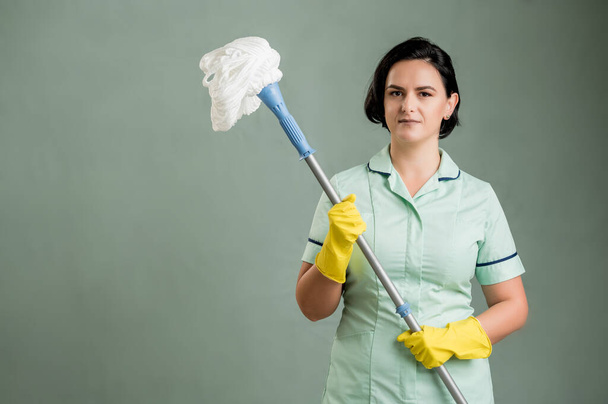Young cleaning woman wearing a green shirt and yellow gloves, holding mop isolated on green background - Photo, Image