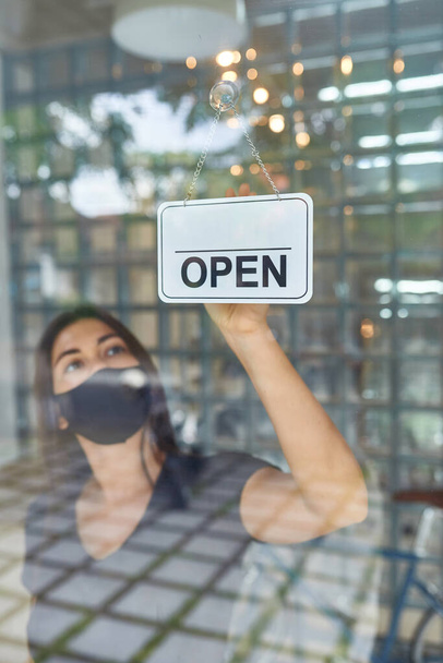 Mooi Europees meisje in een masker hangt een bord op een glazen deur van een kledingzaak (open) na een cowid-19 epidemie - Foto, afbeelding