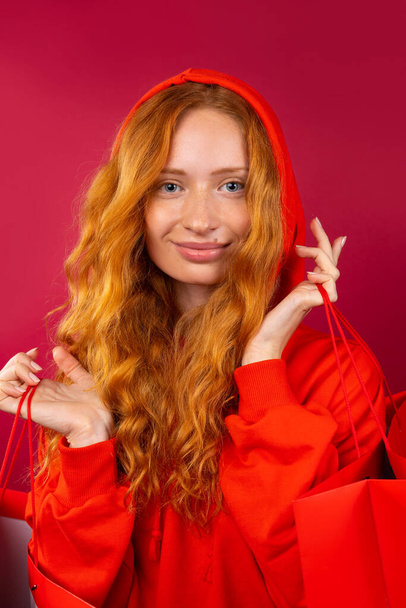 Portrait of a happy young red-haired girl with freckles holding red gift bags on a red background. Holiday and shopping concept - Foto, imagen