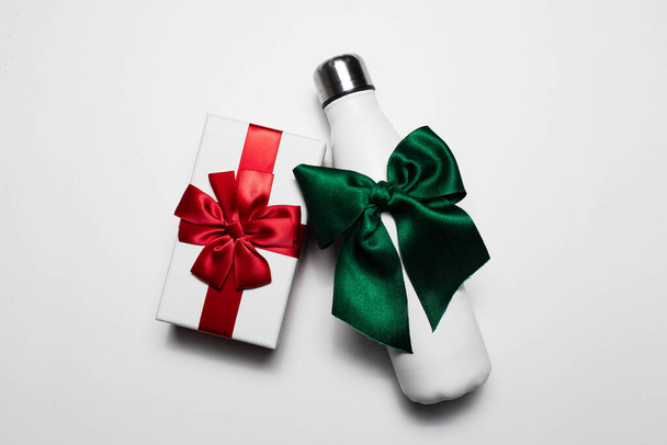 Close-up van geschenkdoos en herbruikbare stalen thermo waterfles met rode en groene strikken. Geïsoleerd op witte achtergrond. - Foto, afbeelding
