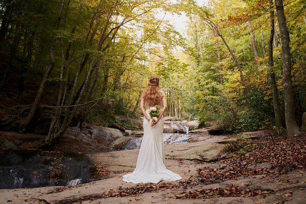 Hochwinkelrückansicht der schönen jungen Braut mit einem Strauß auf dem Rücken im Wald. - Foto, Bild