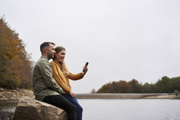 Pareja sentada frente al lago haciendo un selfie al aire libre con un lago en el fondo. - Foto, imagen