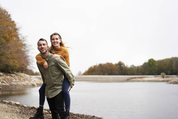 Young man piggibacking his girlfriend and laughing at the lake. - Photo, Image