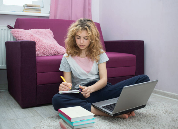 miła dziewczyna - nastolatka w wieku 17 lat, przygotowująca się do egzaminów w domu, siedząca na podłodze przed laptopem, pisząca w notatniku. kształcenie na odległość. Wiedza, książki, nowoczesne technologie cyfrowe - Zdjęcie, obraz