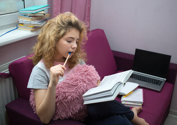 симпатичная девушка - подросток 17 лет, готовится к экзаменам дома, сидит на диване, пишет в тетрадке, думает. дистанционное обучение. Знания, книги, современные цифровые технологии - Фото, изображение