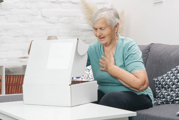  Starší žena otvírá krabici s balíky doma. Šťastný usměvavý starší žena dívá do otevřené balíček krabice doma. - Fotografie, Obrázek