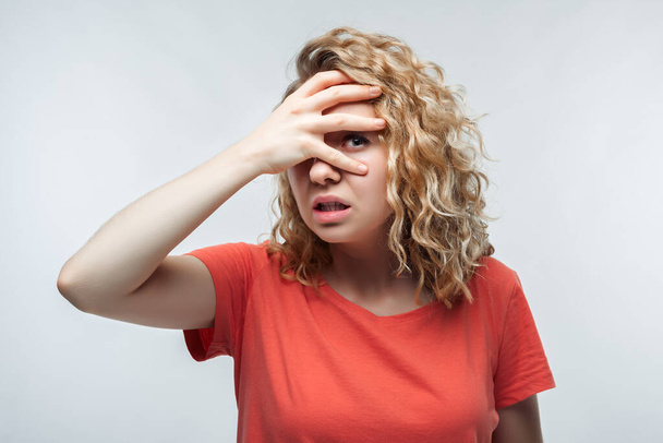 Portrait of frightened young woman with curly hair in casual t shirt covering her eyes with hands. Human emotions, facial expression concept. Studio shot, white background, isolated - Photo, Image