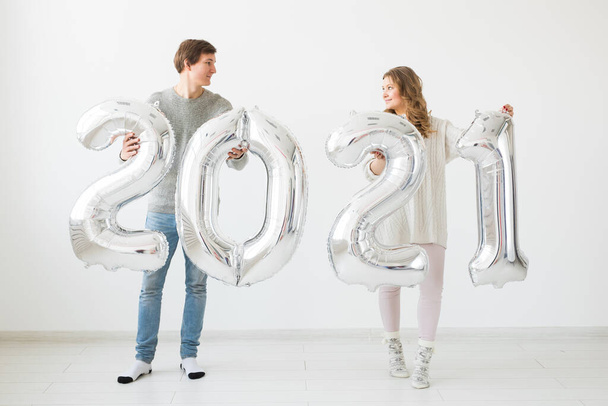 Vacaciones, fiesta y concepto de fiesta - Feliz pareja amorosa sostiene globos de plata 2021 sobre fondo blanco. Celebración de Año Nuevo. - Foto, Imagen