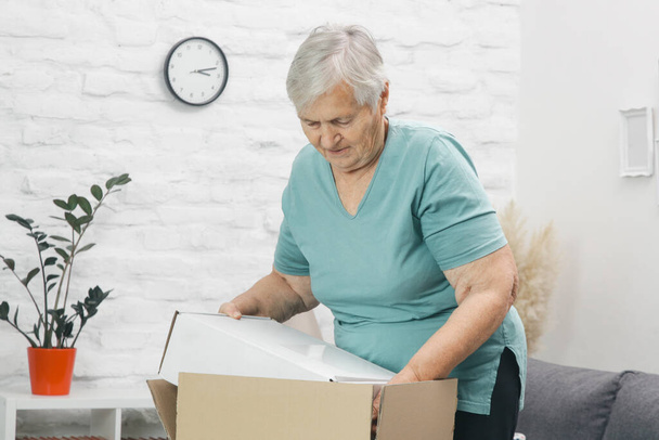 Oudere vrouw opent pakketdoos thuis. Gelukkig glimlachende oudere vrouw op zoek naar open pakket doos thuis. - Foto, afbeelding