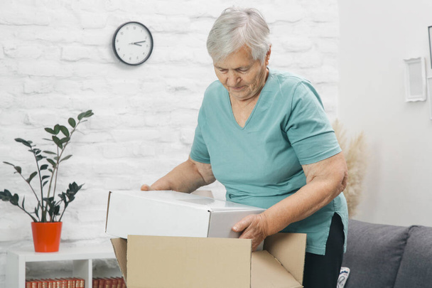 Vanhempi nainen avaamassa pakettilaatikkoa kotona. Onnellinen hymyilevä vanhus nainen etsii avoimeen pakettilaatikkoon kotona. - Valokuva, kuva