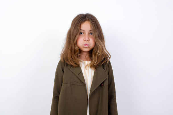 красивая маленькая девочка в зеленом пальто делает грустное лицо на белом фоне   - Фото, изображение
