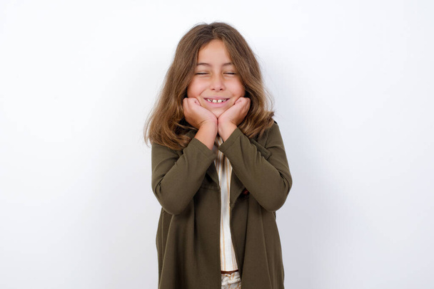 Portret van jong schattig meisje student wordt overweldigd door emoties, uiten van opwinding en geluk met gesloten ogen en handen in de buurt van het gezicht, terwijl glimlachen breed over grijze achtergrond. - Foto, afbeelding
