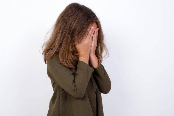 Sad young Caucasian little girl standing against white background wearing casual clothes standing against  background covering his face with hands and crying. - Photo, Image