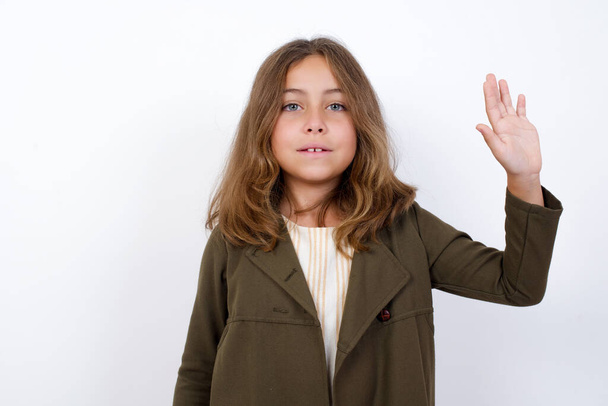 Portrait of cheerful, outgoing friendly-looking young little girl standing against white background  wearing casual clothes raise one hand and wave, saying hi or hello and smiling with carefree expression as make goodbye or welcome gesture. - Foto, Imagem