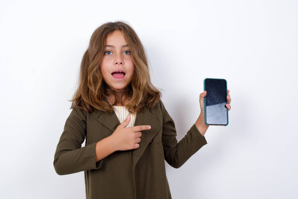 Podekscytowana dziewczynka w zielonym płaszczu wskazująca palcem na urządzenie cyfrowe. Studio shot wstrząśnięty dziewczyna gospodarstwa smartfon z pustym ekranem. - Zdjęcie, obraz
