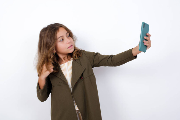 Retrato de una niña muy feliz usando abrigo verde tomando una selfie aislada sobre fondo blanco - Foto, imagen