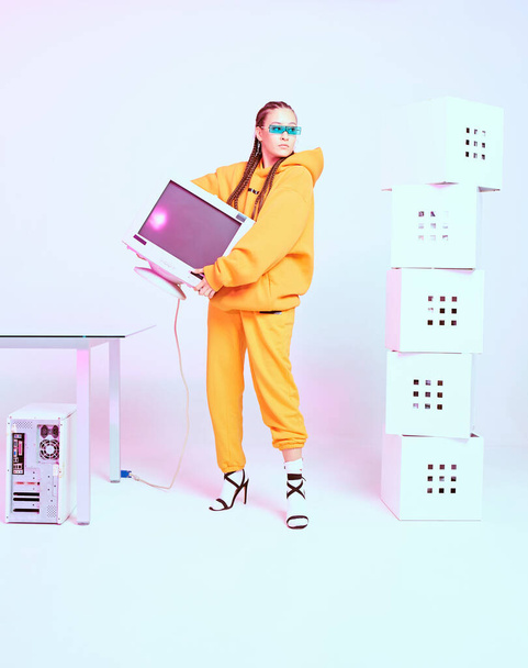 Cool female office worker in sunglasses standing with obsolete computer monitor and boxes. Colored Neon light - Zdjęcie, obraz