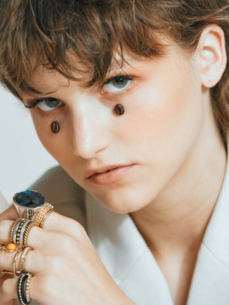 Pretty young girl with coffee beans on face, stylish haircut and rings, close up studio portrait  - 写真・画像