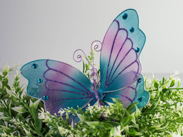 Mariposa grande de malla púrpura y azul con purpurina y gemas encima de una planta blanca y verde con un fondo blanco. Perfecto para primavera! - Foto, Imagen