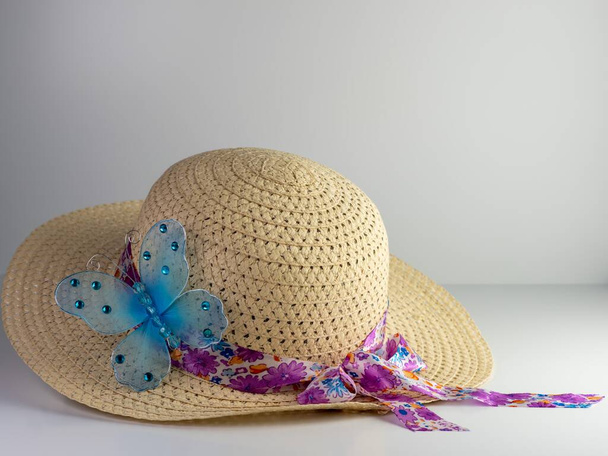 Sombrero de paja con una cinta floral morada y lazo y decorado con una mariposa azul, sobre un fondo blanco. - Foto, imagen