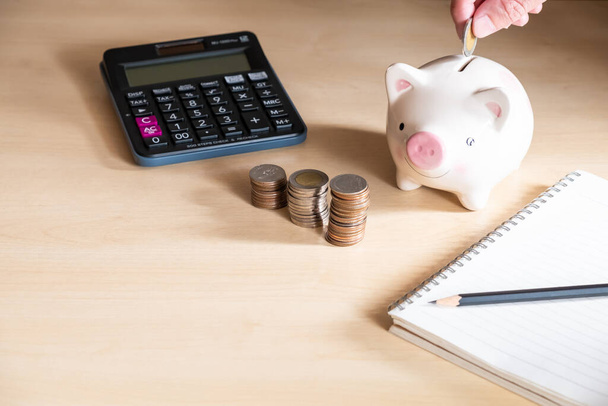 Selective focus at men hand put coin into piggy bank. With blurred calculator and increase stack of money on wooden desk. Personal financial saving concept for retirement or insurance. - Foto, afbeelding