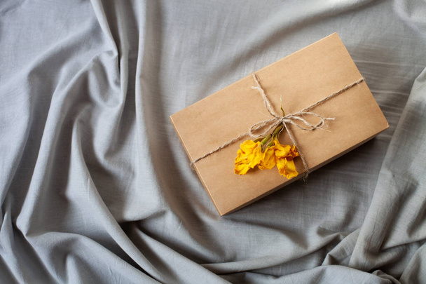 Basit hediye paketi, el işi kutusu, kurdele sicimi, kuru sarı çiçek, gri keten kumaş arka planda. Sıfır atık, üst görünüm. - Fotoğraf, Görsel