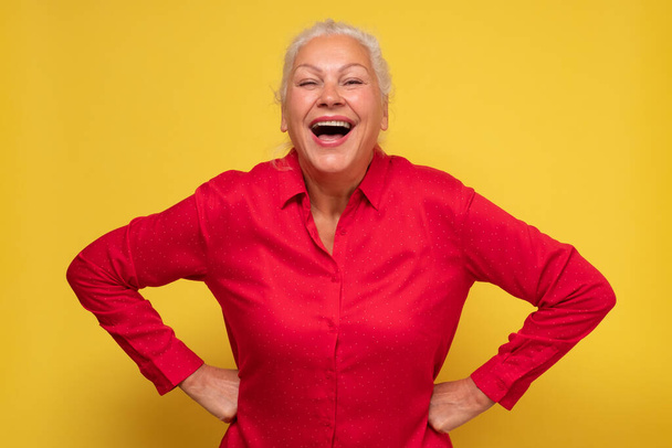 Pewna siebie atrakcyjna starsza kobieta uśmiechnięta szczęśliwie śmiejąca się z kamery na żółtej ścianie. - Zdjęcie, obraz