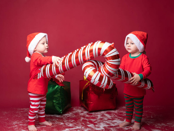 İki yeni yürümeye başlayan erkek kardeş kırmızı Noel Baba şapkalı ikizler ve kırmızı arka planda hediyeler.. - Fotoğraf, Görsel