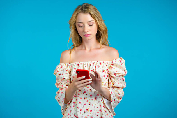 Woman sms texting, using app on smartphone. Pretty girl with blonde hair surfing internet with mobile phone. Blue studio portrait. - Foto, Imagem
