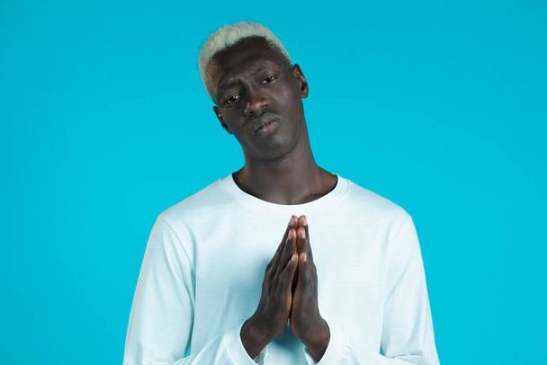Young african man in white long sleeve praying over blue background. Guy begging someone satisfy his desires, help with. - Foto, immagini