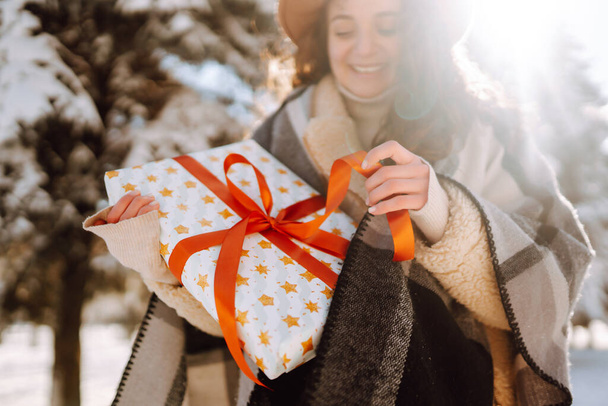 Pretty Woman holding a giftbox with red ribbon in the winter park. Young woman wearing hat, plaid scarf and coat. Happy winter time. Christmas. New years. - Foto, Imagen