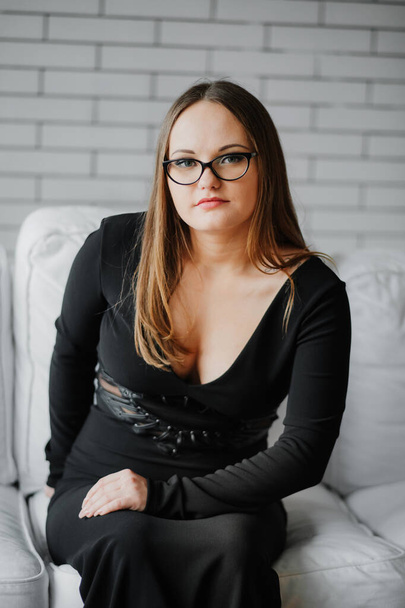 retrato de una joven en gafas y un vestido negro sentado en un sofá blanco. - Foto, Imagen