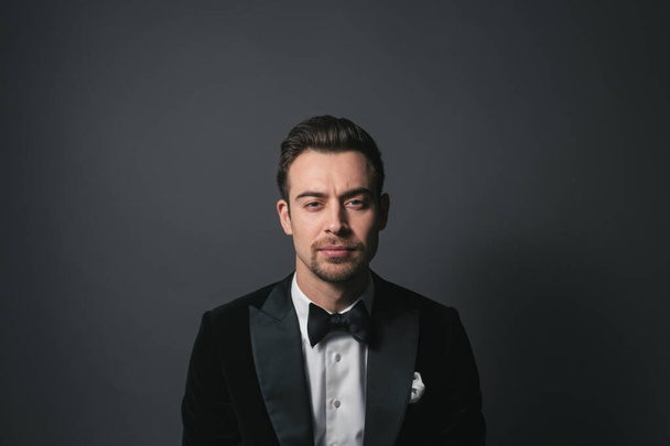 Studio portrait of a young caucasian man in a tuxedo, looking at the camera, against plain studio background - 写真・画像