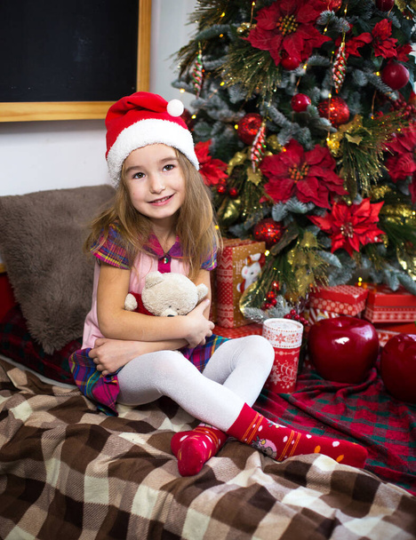 A little girl holding a Teddy bear, sitting on a plaid blanket in the Christmas decorations near a Christmas tree with boxes of gifts and a Santa hat. New year, children's game - Fotoğraf, Görsel