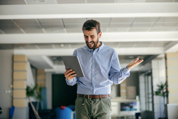 Young smiling bearded businessman walking in hall of his firm and holding tablet. He is overjoyed because he find solution for a problem. - Photo, Image